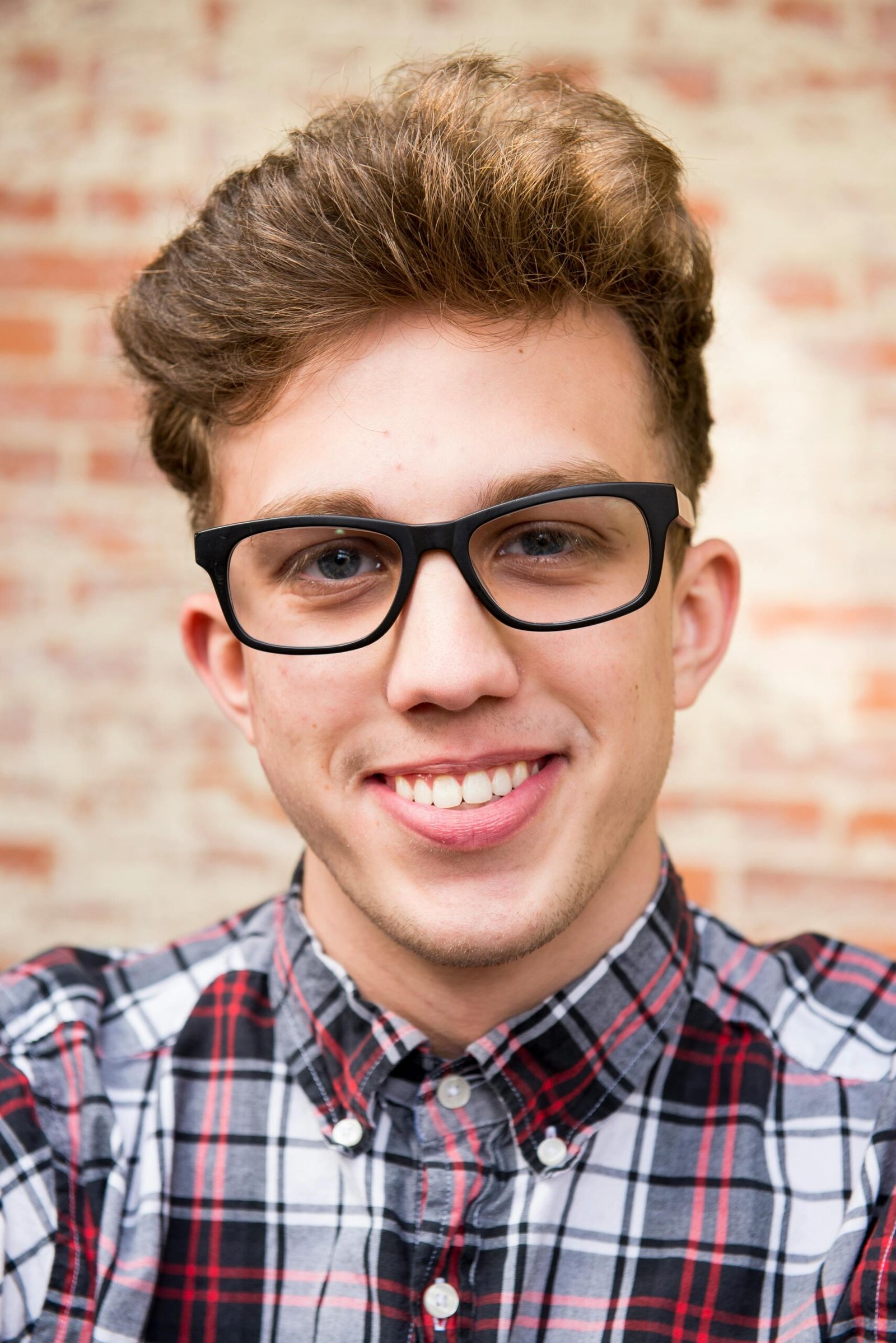 Portrait of a smiling young man wearing eyeglasses and plaid shirt against a brick wall background.